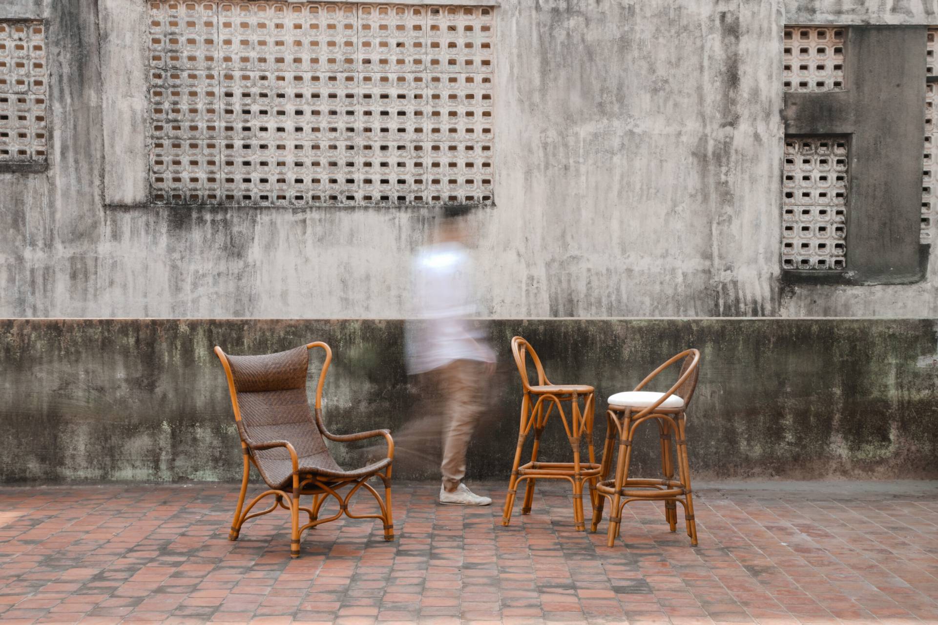 Handcrafted modern cane furniture featuring a rattan lounge chair and two bar stools on a brick patio with a blurred figure walking past.